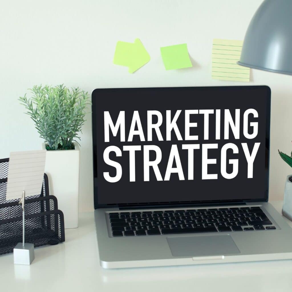 Laptop on a clean desk with the screen reading 'MARKETING STRATEGY' amid a plant and sticky notes.