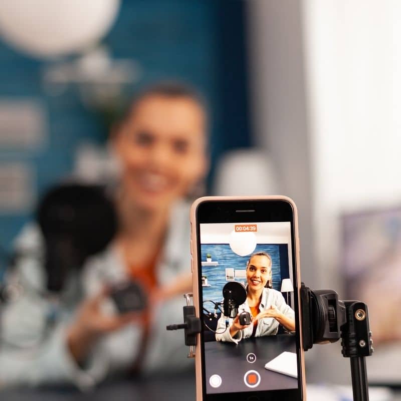 Person filming themselves with a smartphone on a tripod, microphone in front, smiling at the camera (vlog setup)