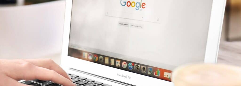 Hands typing on a white MacBook Air with the Google homepage visible on the screen and a cup of coffee nearby on the right.