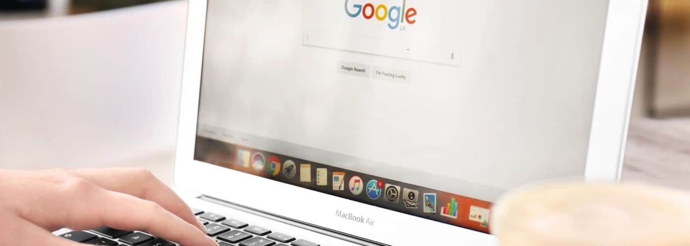 Hands typing on a white MacBook Air with the Google homepage visible on the screen and a cup of coffee nearby on the right.