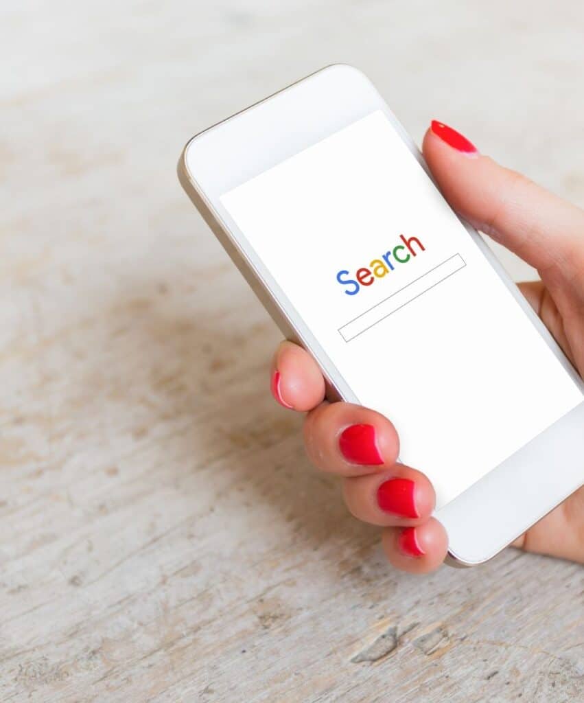 Close-up of a hand with red nails holding a white smartphone displaying a search screen with a colorful 'Search' logo and a search bar.