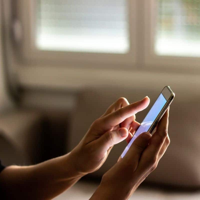 Close-up of hands holding and tapping a smartphone in a bright indoor setting near a window.